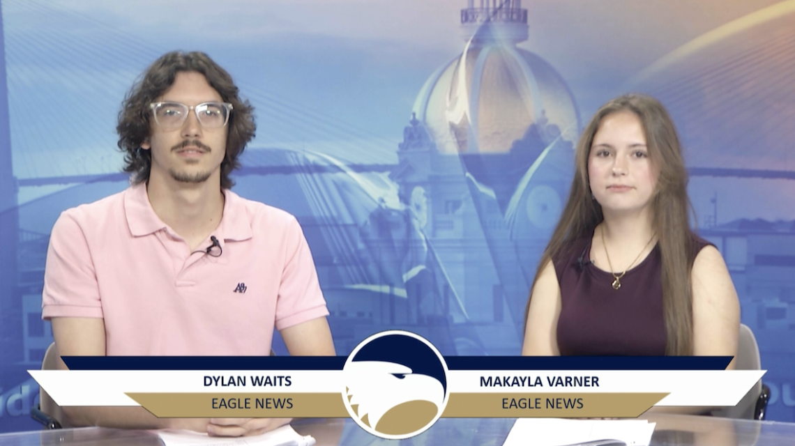 Two student newscasters sit next to each other at a news desk. One has on a pink shirt, and the other has on a maroon shirt.